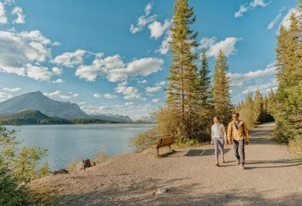 two people walking on a trail next to Kananaskis Lake in the summer. There are blue skies with clouds and mountains in the background.