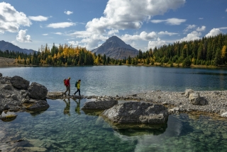 two people walking along a lake with fall colours and mountains in the background