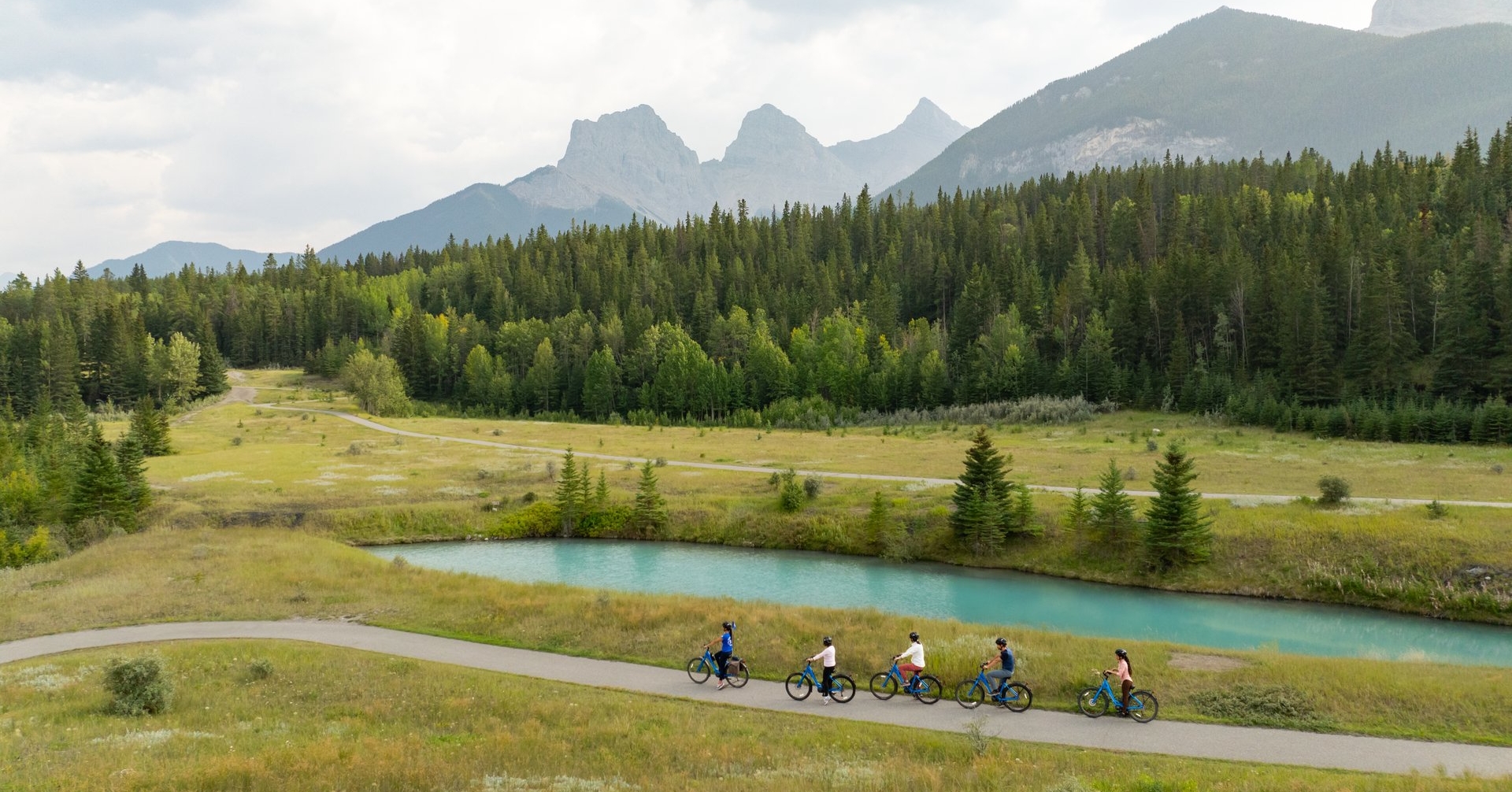 a food tour group cycling through canmore next to a pond with the three sisters mountains in the background