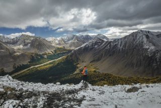 a person standing on top of a mountain ridge with snow looking down on the fall colours in the valley below