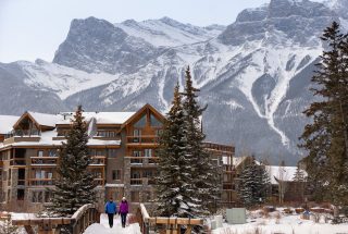two people walking across a bring in spring creek next to the malcolm hotel with snow covered mountains in the background