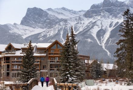 two people walking across a bring in spring creek next to the malcolm hotel with snow covered mountains in the background