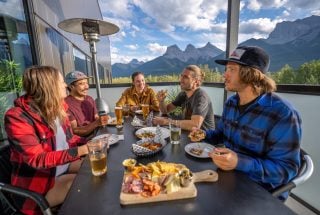 a group of people eating food and drinks on the canGOLF outdoor patio in the summer with the Three Sisters Mountains in the background