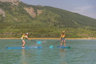 two women splashing each other on stand-up paddleboards on Barrier Lake in the summer with mountains in the background