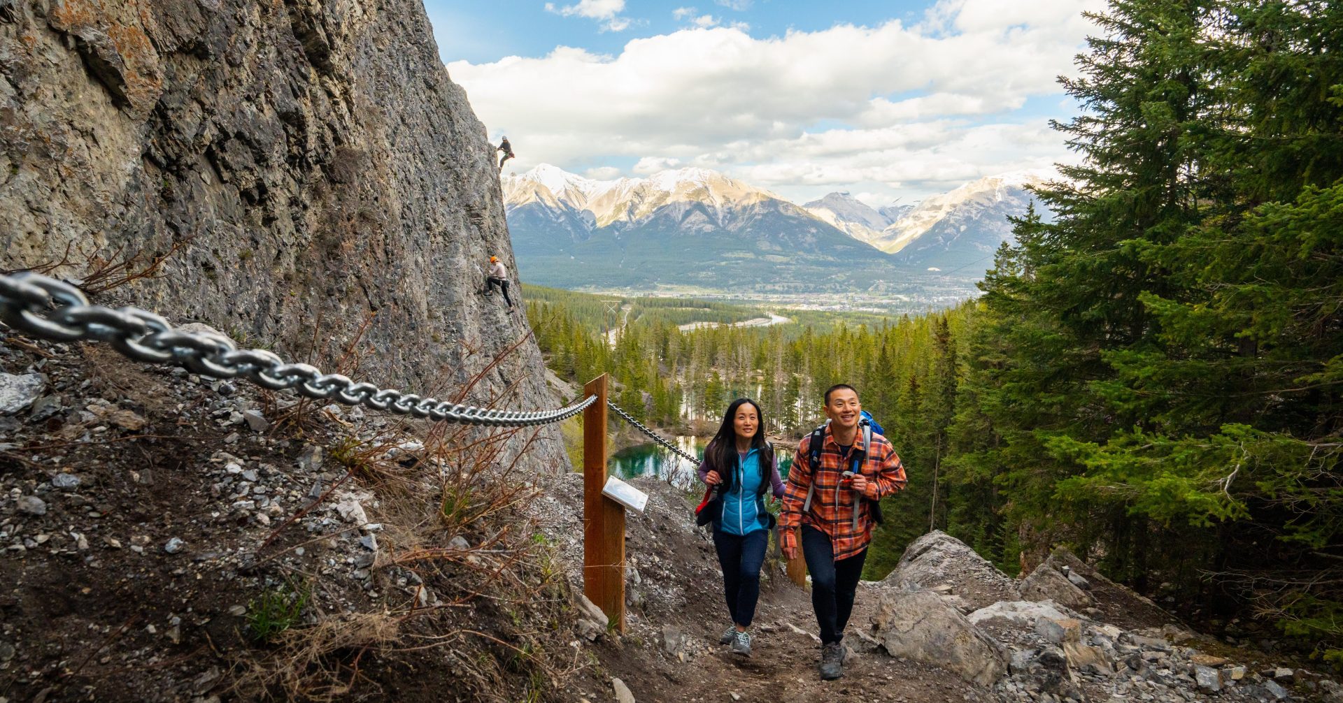 a couple hiking up Grassi Lakes Trail with rock climbers climbing the mountain in the background
