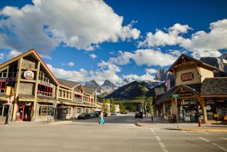 a woman walking on a crosswalk between buildings on Canmore's Main Street on a sunny day with the Three Sisters Mountains in the background