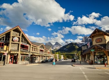 a woman walking on a crosswalk between buildings on Canmore's Main Street on a sunny day with the Three Sisters Mountains in the background