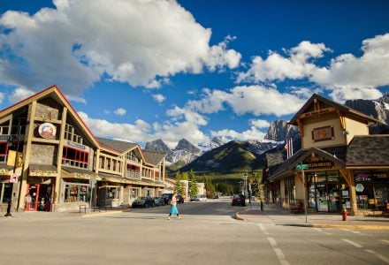 a woman walking on a crosswalk between buildings on Canmore's Main Street on a sunny day with the Three Sisters Mountains in the background