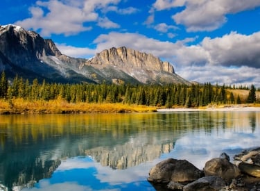 a landscape photo of mount yamnuska in the fall with a reflection in a pond in the foreground and blue skies with scattered clouds