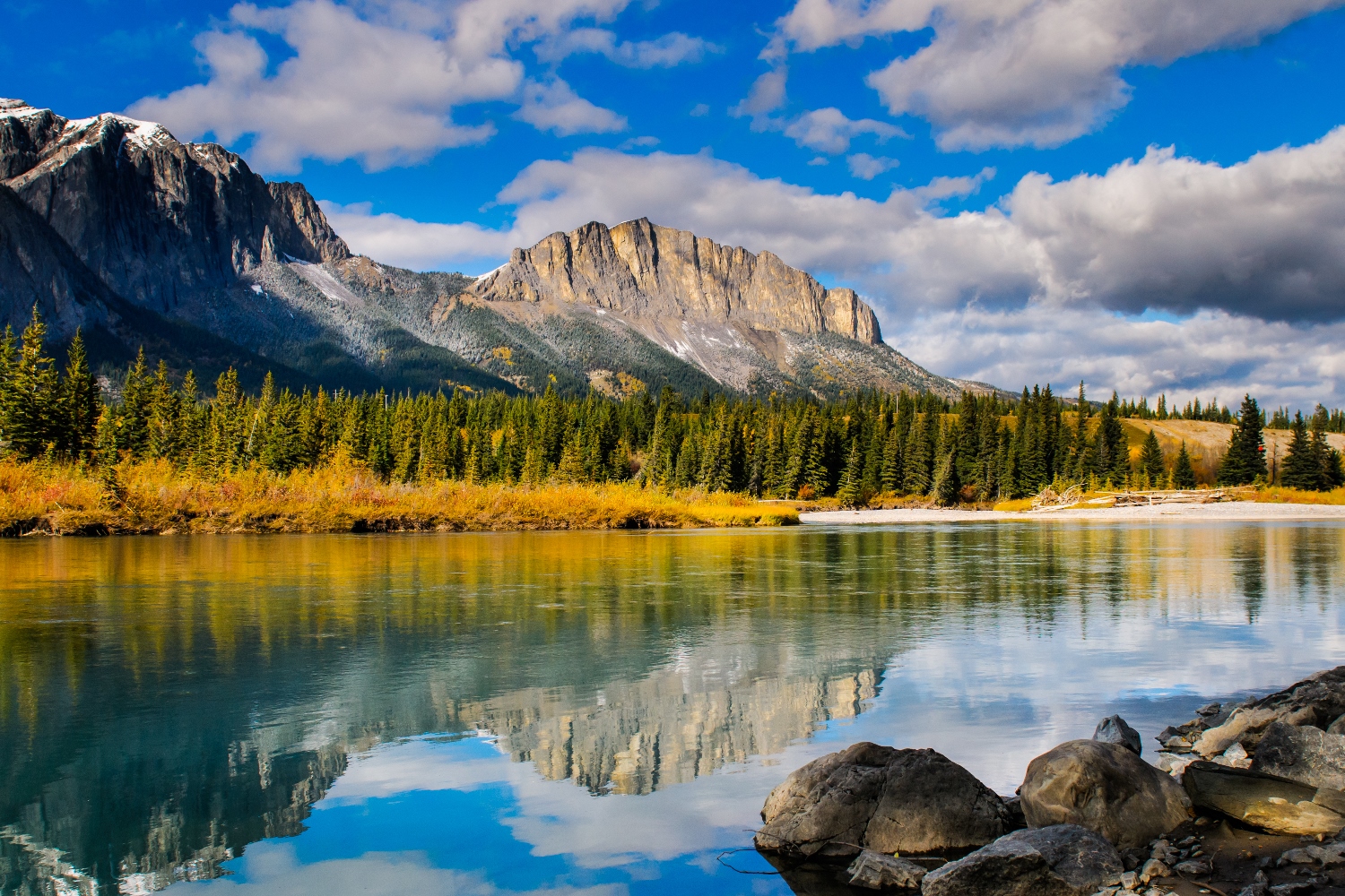 a landscape photo of mount yamnuska in the fall with a reflection in a pond in the foreground and blue skies with scattered clouds
