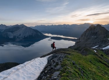 a solo hiker at the peak of a mountain with alpine lakes and mountain ranges in the background. Photo was taken at sunrise