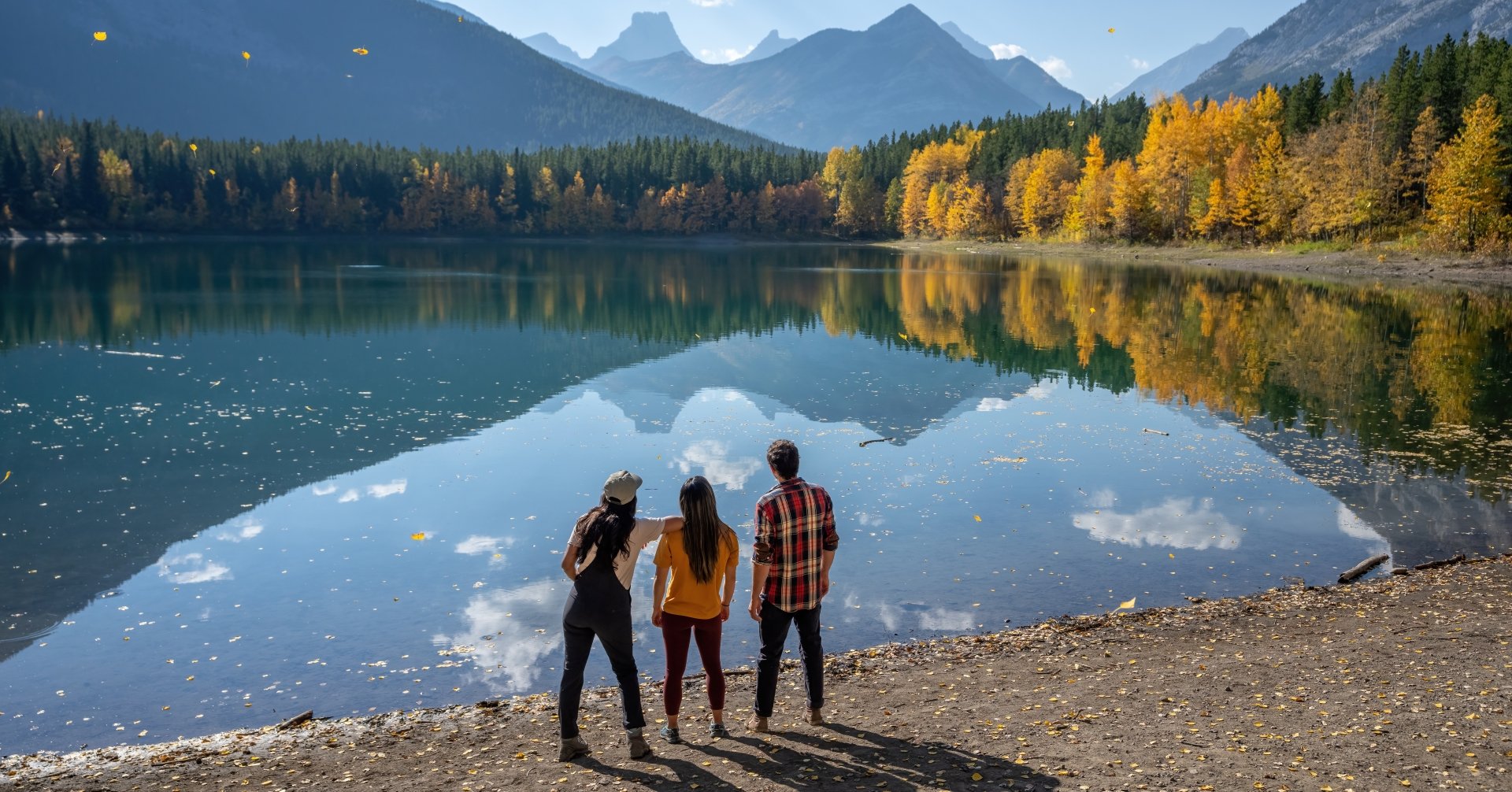 three people standing in front of a reflective pond with fall colours and mountains lining the pond