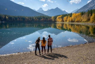 three people standing in front of a reflective pond with fall colours and mountains lining the pond