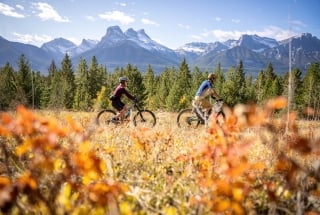 two bikers on a mountain bike trail with fall colours in the foreground and the snow capped three sisters mountains in the background