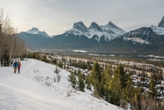 Two pedestrians walking along a snowy path along Benchlands with the town of Canmore below and the Three Sisters Mountains in the background