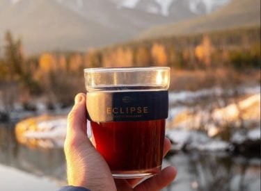 a person holding up a cup of eclipse coffee with the three sisters mountains and fall colours in the background