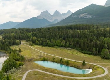 an aerial photo of cyclists on the west bow river pathway with the three sisters mountains in the background and a blue runoff pond